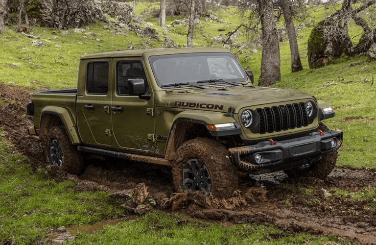 2025 Jeep Gladiator drives through a muddy off-road trail surrounded by lush greenery and rocky terrain.