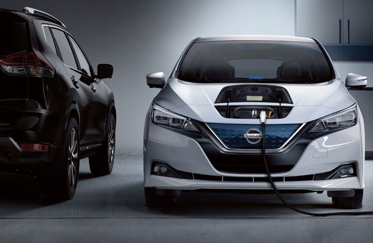 White 2019 Nissan Leaf with a compartment on its hood opened up and a big charging cord plugged in. Head-on view parked in a garage by a cabinet.