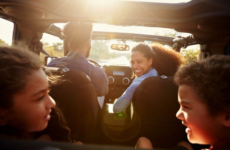 An African American mother seated in the passenger's seat, is smiling at the children in the backseat while her husband drivers the SUV