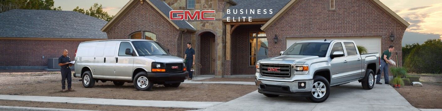 GMC Business Elite banner with Savana and Sierra models parked on a driveway outside a house garage as workers inspect the models