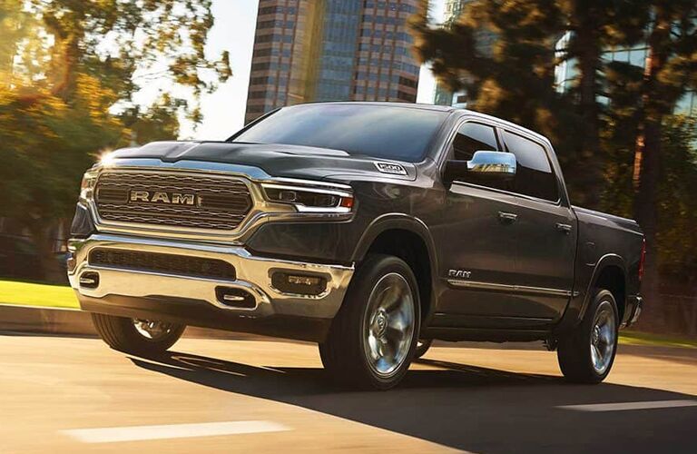 2019 Ram 1500 exterior shot with gray paint color driving through a city road near trees and a skyscraper in the background as the sun shines