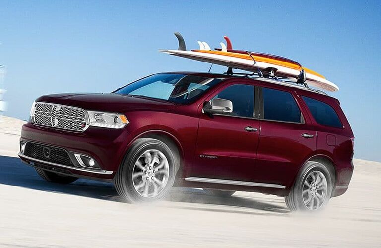 2019 Dodge Durango exterior side shot with red purple paint color driving on a sand dune and kicking up dust with surf boards on its roof rack rails